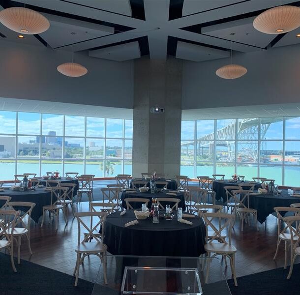 A modern event space with round tables and wooden chairs, set for dining. Large windows offer a view of a river, city buildings, and a bridge under a partly cloudy sky.