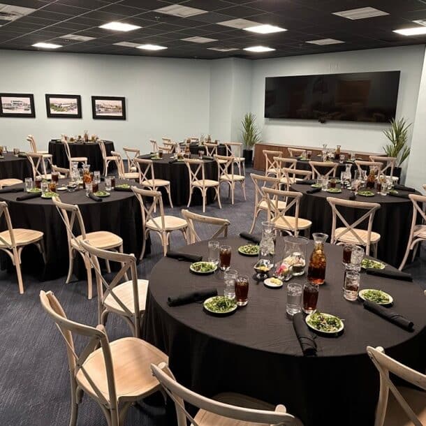 A banquet room with several round tables covered in black tablecloths, set with drinks, salads, and neatly folded black napkins. Wooden chairs are arranged around the tables, and framed photos hang on the walls.