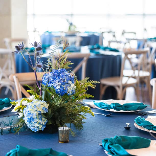A round table set for an event features teal napkins, white plates, and a centerpiece of blue hydrangeas and greenery. Other similarly decorated tables are visible in the bright, sunlit background.